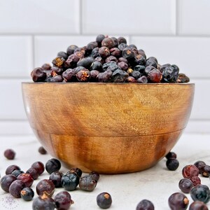 May include: A wooden bowl filled with dried juniper berries. The berries are dark purple and black in color. Some berries are scattered on a white surface.
