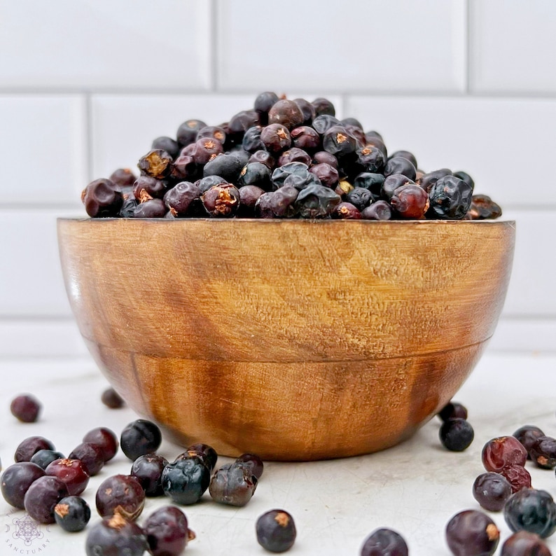 May include: A wooden bowl filled with dried juniper berries. The berries are dark purple and black in color. Some berries are scattered on a white surface.
