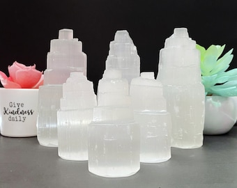 May include: A set of seven white selenite crystal towers of varying sizes. The crystals are smooth and translucent, with a layered, striated texture. The crystals are arranged in a semi-circle on a gray surface.