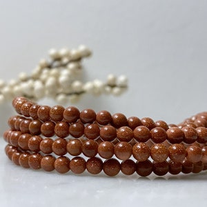 May include: A strand of brown sandstone beads, with a speckled texture, arranged in a circular pattern.