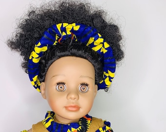Puede incluir: Una muñeca con un vestido estampado en azul, amarillo y rojo con una diadema a juego. La muñeca tiene ojos marrones y cabello oscuro y rizado.