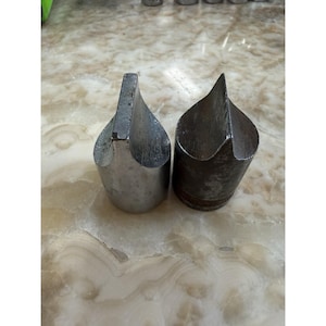 May include: Two metal tools with cylindrical bases and pointed, wedge-shaped tops. The left tool has a smooth, polished silver finish, while the right tool features a darker, textured, matte grey surface with a knurled band near the bottom edge.