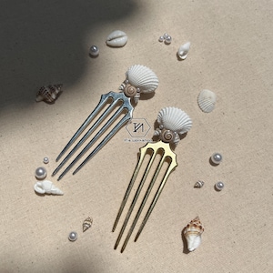 Könnte beinhalten: Zwei Haarkämme mit Muschel-Design. Die Kämme sind golden und silbern und haben drei Zinken. Die Muscheln sind weiß und haben ein kleines Schnecken-Design.