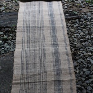 May include: A long, narrow, woven rug with a striped pattern. The rug is made of natural fibers and has a rustic, vintage look. The stripes are a dark gray color on a light beige background.