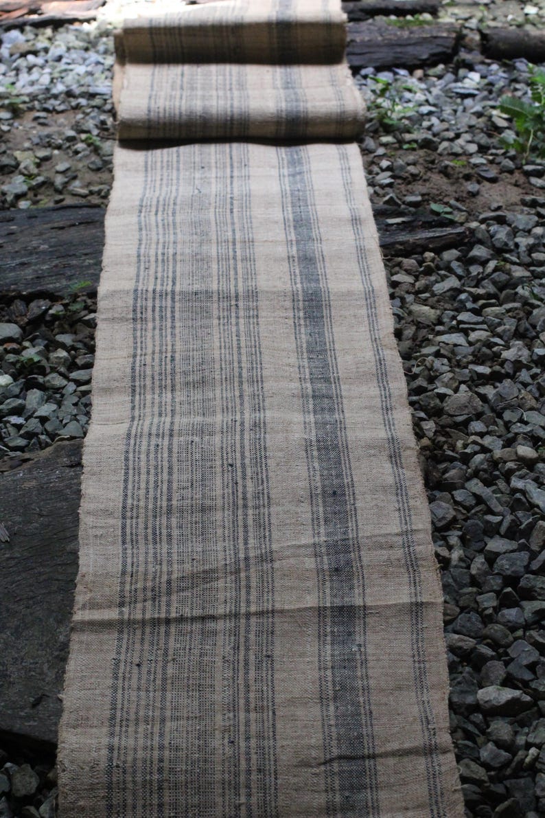 May include: A long, narrow, woven rug with a striped pattern. The rug is made of natural fibers and has a rustic, vintage look. The stripes are a dark gray color on a light beige background.