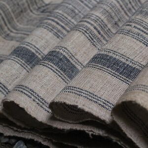 May include: Close-up of a stack of fabric with a striped pattern. The fabric is a light beige color with dark blue stripes. The fabric is folded and layered, creating a textured look.