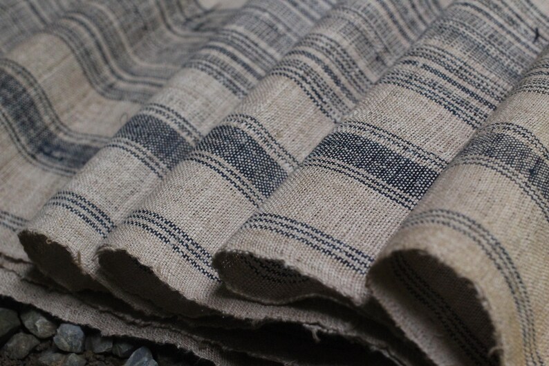 May include: Close-up of a stack of fabric with a striped pattern. The fabric is a light beige color with dark blue stripes. The fabric is folded and layered, creating a textured look.