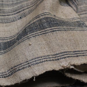 May include: Close-up of a woven fabric with a striped pattern. The fabric is a light beige color with dark blue stripes. The fabric is textured and has a rough feel.