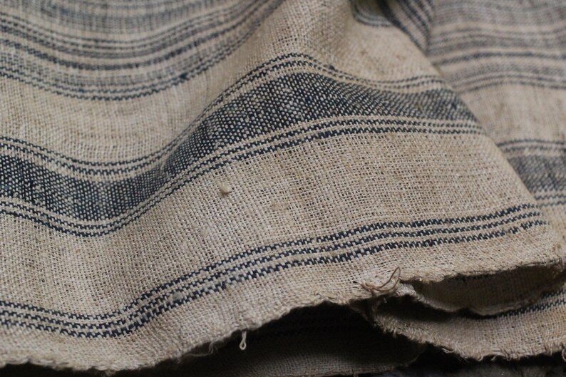 May include: Close-up of a woven fabric with a striped pattern. The fabric is a light beige color with dark blue stripes. The fabric is textured and has a rough feel.