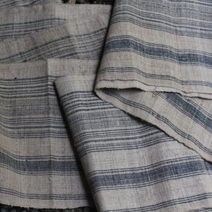 May include: A close-up of a stack of fabric with a striped pattern. The fabric is a light beige color with dark blue stripes. The fabric is woven and has a rough texture.