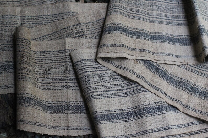 May include: A close-up of a stack of fabric with a striped pattern. The fabric is a light beige color with dark blue stripes. The fabric is woven and has a rough texture.