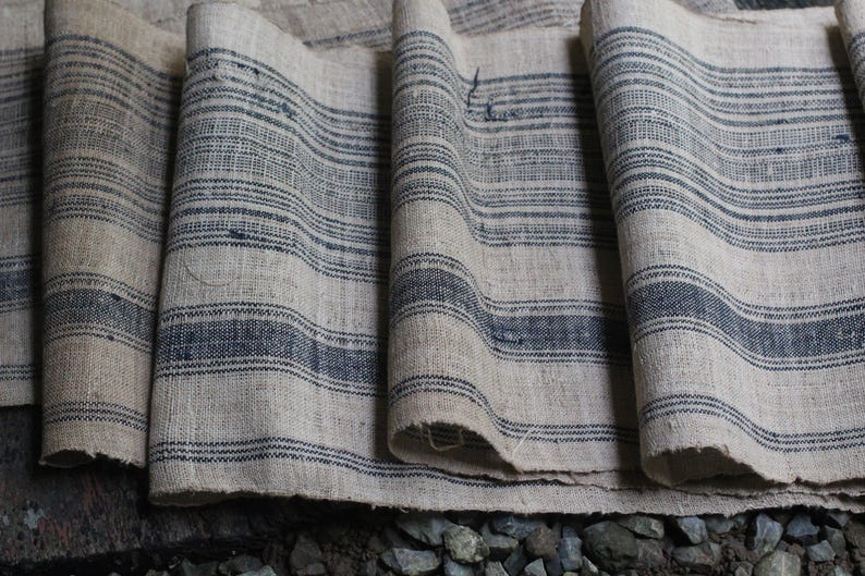 May include: A close-up of a stack of fabric with a striped pattern. The fabric is a light beige color with dark blue stripes. The fabric is folded and layered, creating a textured look.