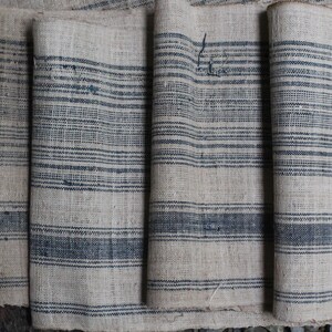 May include: Close-up of a stack of fabric with a striped pattern. The fabric is a light beige color with dark blue stripes. The fabric is woven and has a rough texture.