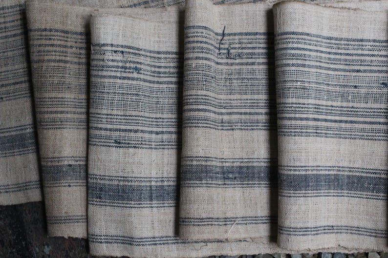 May include: Close-up of a stack of fabric with a striped pattern. The fabric is a light beige color with dark blue stripes. The fabric is woven and has a rough texture.