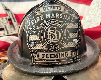 May include: A black fire helmet featuring a front shield with the text DEPUTY FIRE MARSHAL, STAFFORD FIRE DEPT, 1947, and FLEMING. The shield displays a central emblem with the letters FSD and a stylized American flag background pattern.