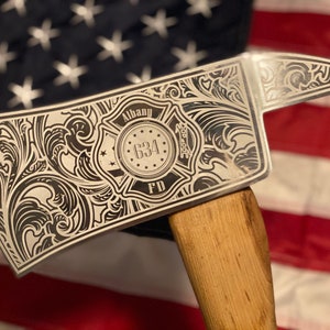 May include: A silver fire axe with intricate floral and scrollwork engraving. The axe head features the text "Albany FD 634" within a shield design. The wooden handle is visible, and the background is an American flag.
