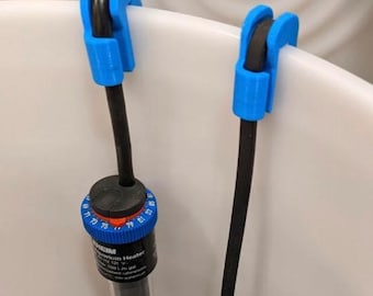 以下が含まれることがあります： 白い容器の縁に取り付けられた2つの明るい青色のプラスチック製クリップが、黒い電源コードとEheim製水槽用ヒーターを固定しています。ヒーターには温度目盛りが付いた青いダイヤルと、黒い円筒形の本体があります。