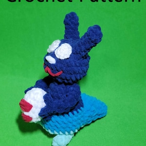 May include: Crochet pattern for a blue and white amigurumi toy. The toy is shaped like a rabbit with a large head and long ears. The toy is holding a white ball with a red stripe.