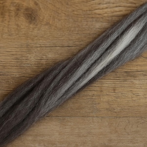 May include: A bundle of gray and white fiber, likely wool or a similar material, is displayed diagonally against a wooden background. The fiber transitions from dark gray to lighter shades, with a central white stripe. The texture appears soft and slightly fluffy.