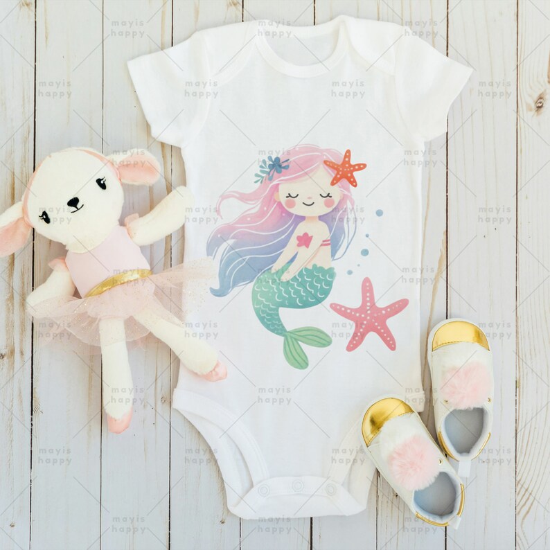 May include: A white baby onesie with a mermaid illustration. The mermaid has pink hair, a green tail, and is holding a red starfish. The onesie is on a white wooden background.