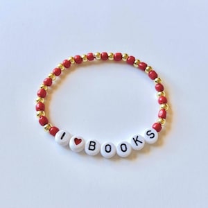 May include: A beaded bracelet with red and gold beads. The bracelet spells out "I ❤️ BOOKS" using white letter beads and a red heart bead. The bracelet is on a white surface.