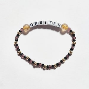 May include: A beaded bracelet featuring white square letter beads spelling ORBITER, flanked by two translucent round beads with gold star accents. The band consists of alternating small black round beads, metallic gold spheres, and faceted purple crystals.