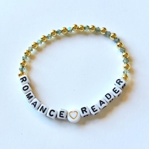 Puede incluir: Pulsera de cuentas con las palabras "ROMANCE READER" escritas con cuentas blancas. La pulsera presenta cuentas doradas y pequeñas cuentas transparentes de color azul verdoso. Una cuenta blanca con un corazón dorado separa las palabras.