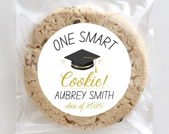 May include: A chocolate chip cookie in a clear cellophane bag. The cookie has a white circular sticker with the text "ONE SMART Cookie!" and a graduation cap graphic. The sticker also includes the name "AUBREY SMITH" and "class of 2026".