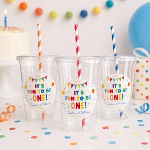 Puede incluir: Vasos de plástico transparente con pajitas a rayas y etiquetas que dicen "IT'S FUN TO BE ONE!" para una fiesta de primer cumpleaños. Las pajitas tienen rayas naranjas, rojas y azules. Confeti y una tarta con una vela encendida.
