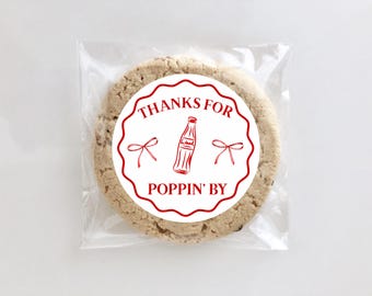 Puede incluir: Una galleta con chispas de chocolate en una bolsa de celofán transparente. La galleta tiene una pegatina redonda roja y blanca con las palabras "THANKS FOR POPPIN' BY" y una ilustración de una botella de refresco.