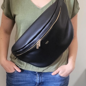 May include: Black leather fanny pack with a gold zipper and hardware. The bag is worn across the body. The word "MYA" is printed on the front of the bag. The person is wearing a green shirt and blue jeans.