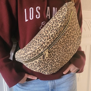 May include: A leopard print fanny pack with a gold zipper and hardware. The bag is worn across the body. The person is wearing a maroon hoodie with the text "LOS AN" in white.