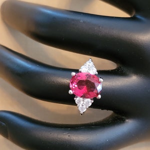 May include: A silver ring with a large oval-shaped red gemstone surrounded by smaller clear gemstones.