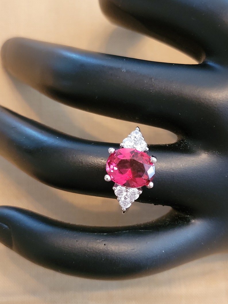 May include: A silver ring with a large oval-shaped red gemstone surrounded by smaller clear gemstones.