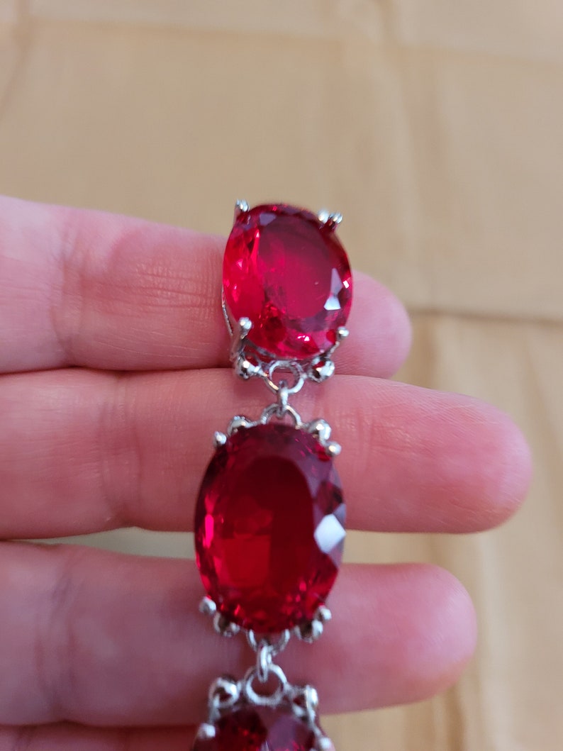 Puede incluir: Un collar de cadena de plata con tres piedras preciosas rojas grandes de forma ovalada. Las piedras preciosas est&aacute;n engastadas en plata.