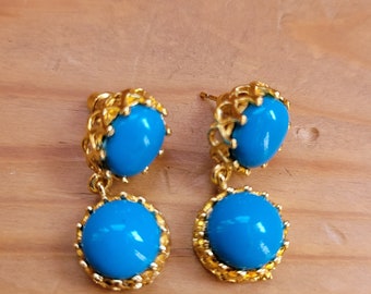Puede incluir: Un par de pendientes colgantes de tono dorado con piedras ovaladas azules. Los pendientes tienen un diseño delicado y adornado.