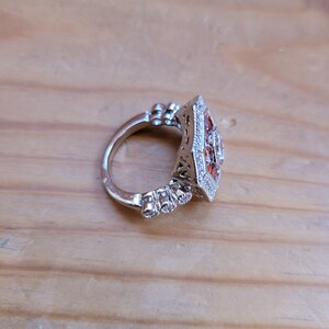 May include: A silver ring with a square-shaped setting featuring a cluster of orange and clear gemstones. The ring has a delicate, ornate design.