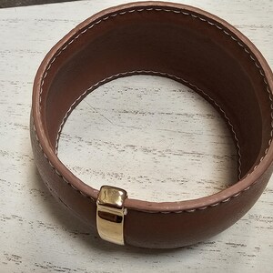 May include: A brown leather bracelet with a gold metal clasp. The bracelet is stitched with white thread.