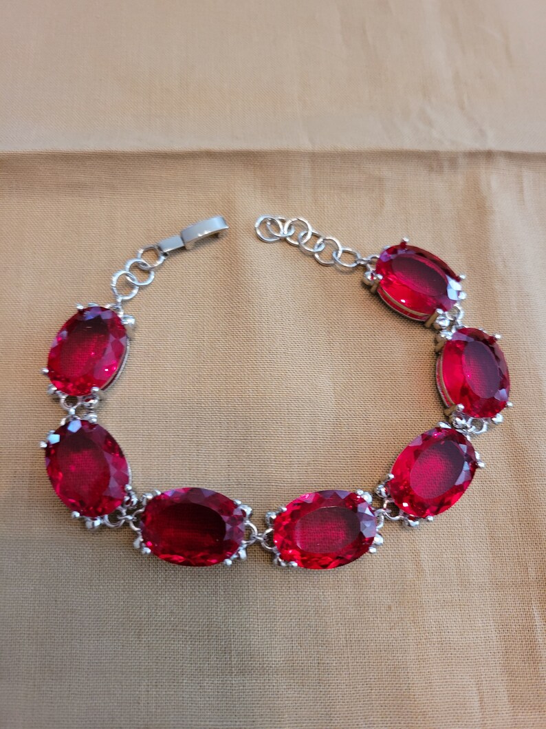 Puede incluir: Un brazalete de plata con un dise&ntilde;o de cadena y piedras preciosas rojas de forma ovalada.