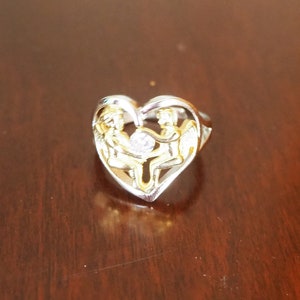 May include: A silver heart-shaped ring with two gold angels holding a clear crystal in the center.