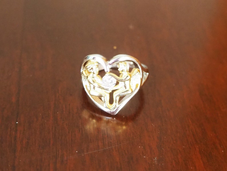 May include: A silver heart-shaped ring with two gold angels holding a clear crystal in the center.