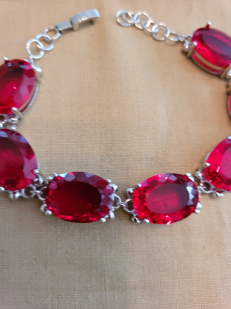 Puede incluir: Un brazalete de plata con piedras preciosas ovaladas rojas. El brazalete tiene un cierre de broche.