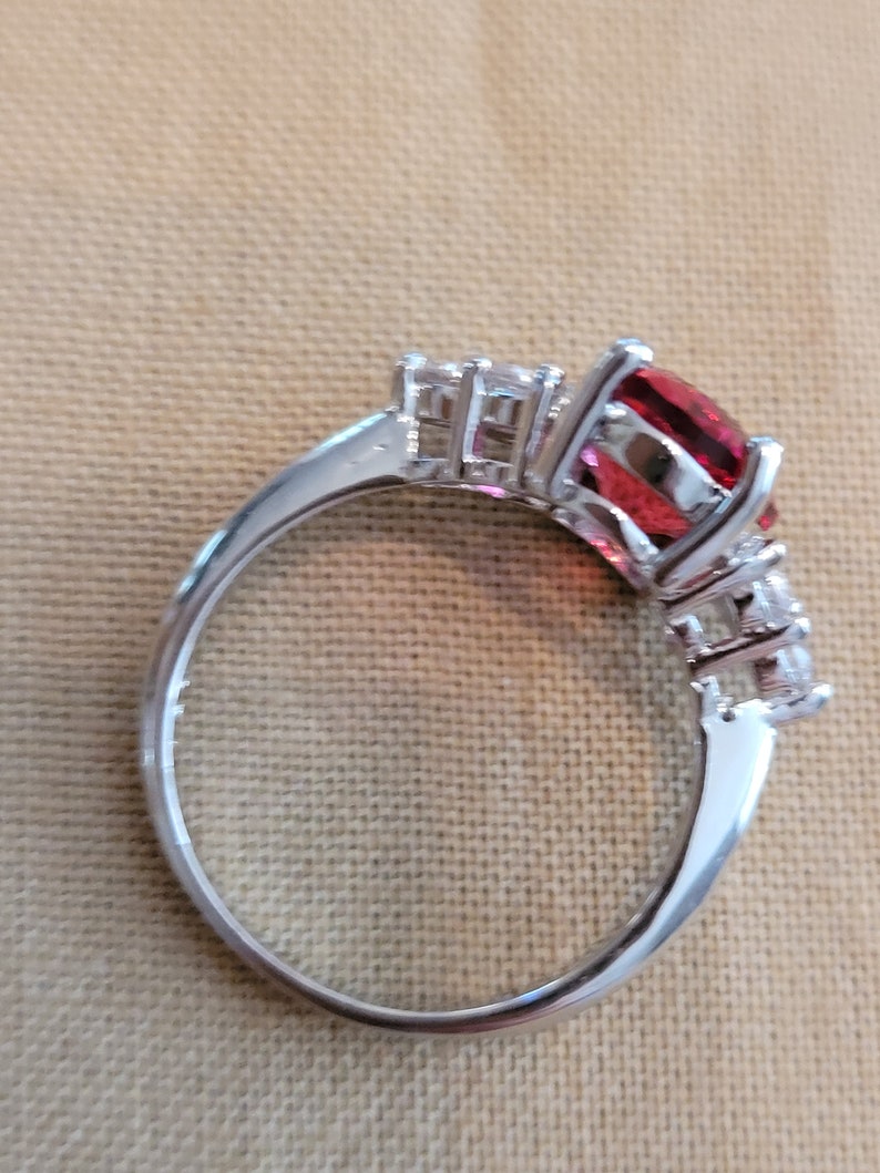 May include: A silver ring with a large red gemstone and smaller clear gemstones on the band.