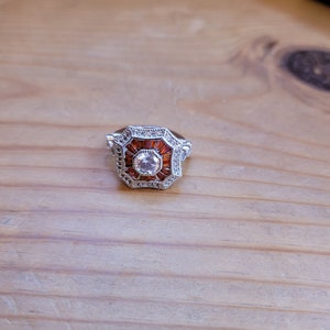 May include: A silver ring with a large, octagonal-shaped stone set in the center. The stone is surrounded by smaller, clear stones. The ring has a vintage style.