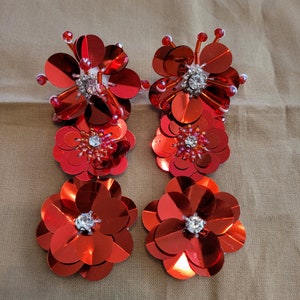 Può includere: Un paio di orecchini rossi a forma di fiore con decorazioni scintillanti. Ogni orecchino presenta tre strati di paillettes rosse a forma di fiore, con un cristallo trasparente al centro di ogni fiore.