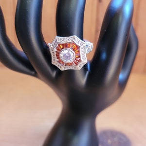 May include: A silver ring with a large octagonal center stone surrounded by smaller orange stones and a halo of smaller clear stones.