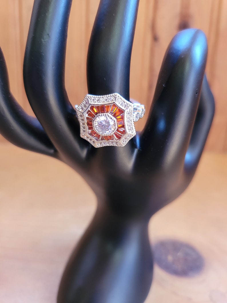 May include: A silver ring with a large octagonal center stone surrounded by smaller orange stones and a halo of smaller clear stones.