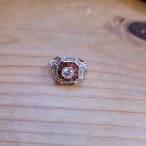 May include: A silver ring with a large, clear, round gemstone surrounded by smaller, orange gemstones. The ring has a geometric, octagonal design.