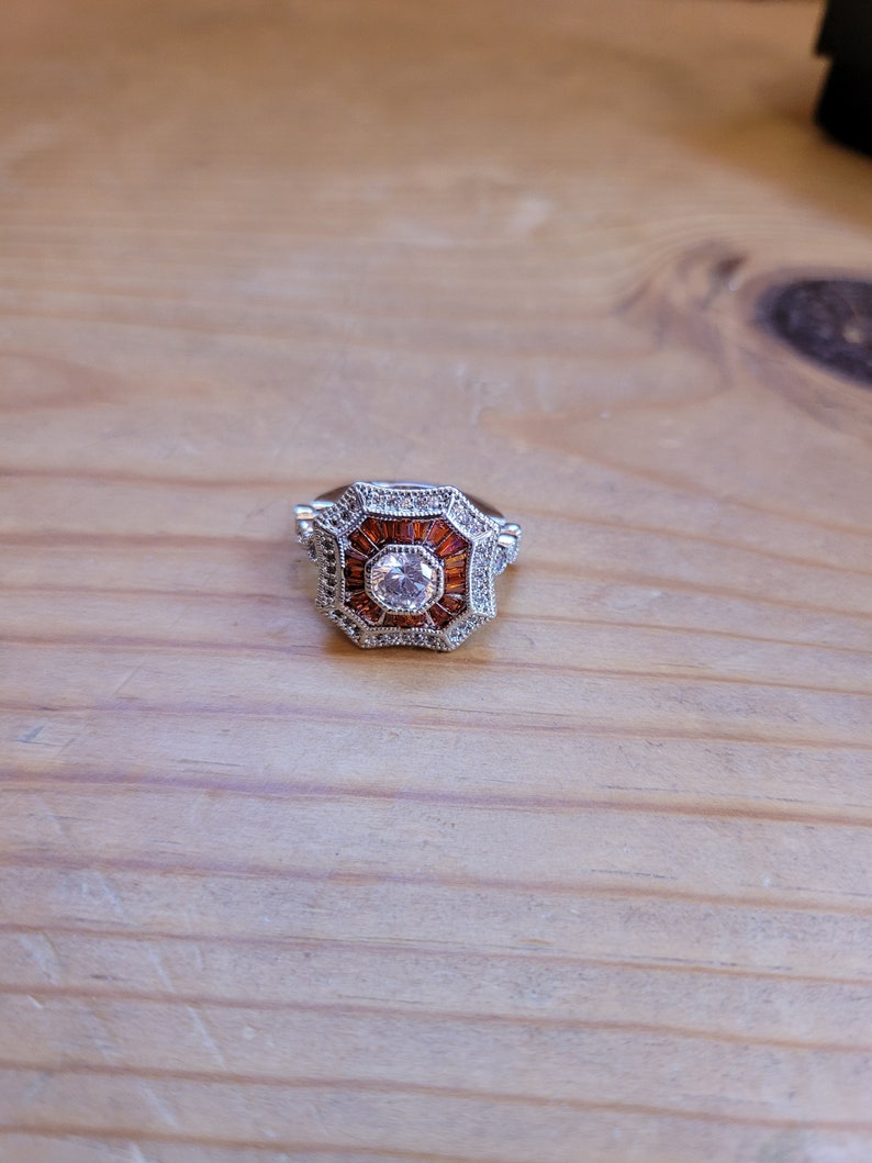 May include: A silver ring with a large, clear, round gemstone surrounded by smaller, orange gemstones. The ring has a geometric, octagonal design.