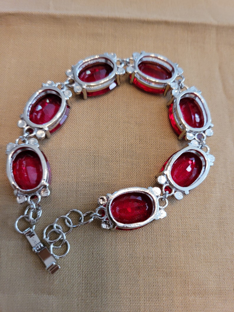 Puede incluir: Un brazalete de plata con cierre de cadena y siete piedras preciosas rojas de forma ovalada.